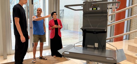 En la imagen se ve a Alfredo, técnico de deportes, a los pies de la escalera del Hall explicando a Ander el funcionamiento de la plataforma elevadora. Marta, la alcaldesa, está presente también. La plataforma está colocada para iniciar el ascenso